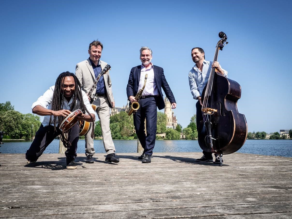 Vier Musiker mit ihren Instrumenten posieren auf einem Holzsteg vor einem See.