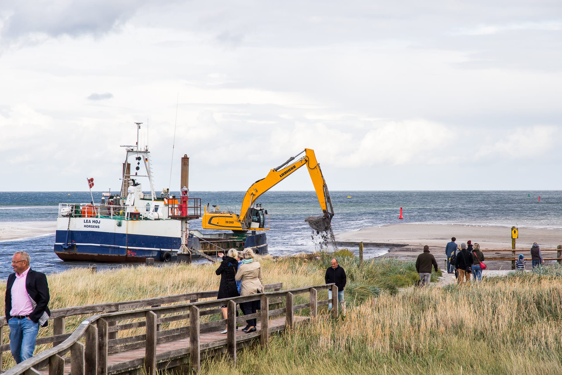 Eine Sandaufschüttung am Strand, bei der ein gelber Bagger Sand ins Wasser kippt. Im Vordergrund gehen mehrere Menschen auf einem Holzweg entlang, und im Hintergrund ein Schiff auf dem Wasser.