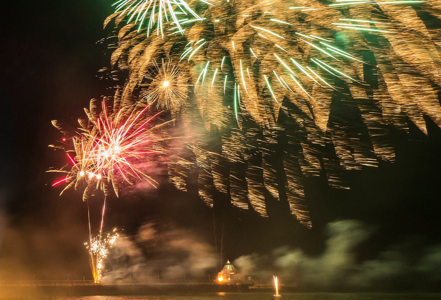 Ein großes, farbenfrohes Feuerwerk explodiert nachts über einer beleuchteten Seebrücke am Meer.