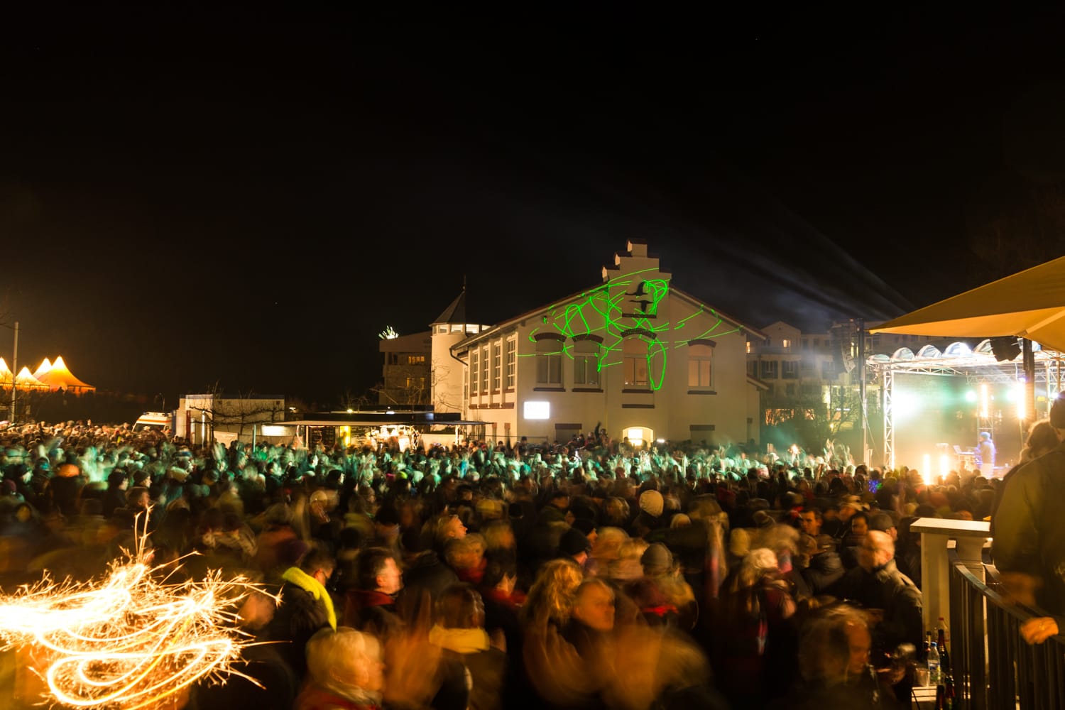Eine große Menschenmenge feiert nachts im Freien vor einem weißen Gebäude mit einem projizierten grünen Symbol und einem Feuerwerk links.