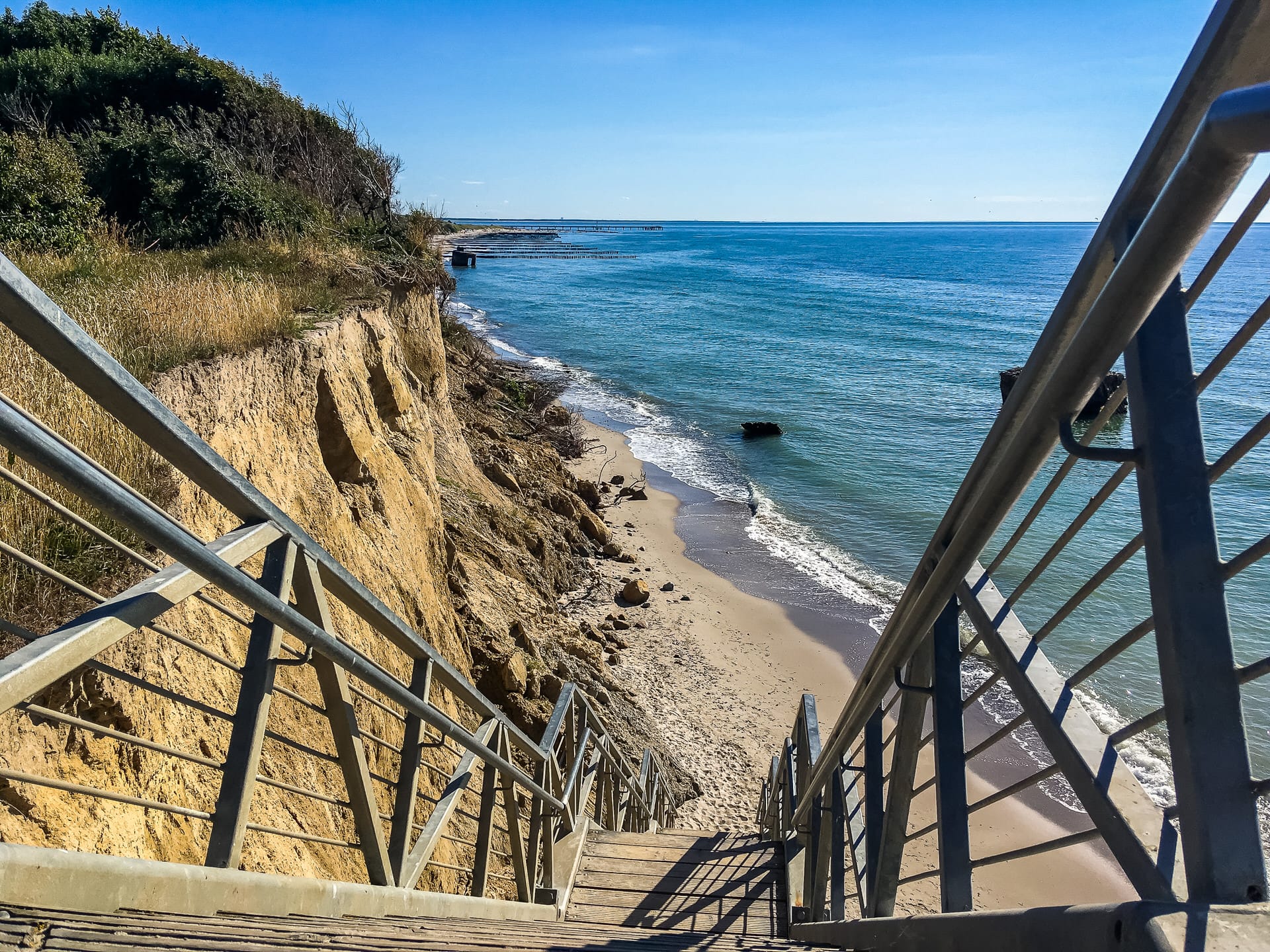 Blick von der Treppe hinunter von der Steilküste auf den sandigen Strand und das Meer, blauer Himmel.