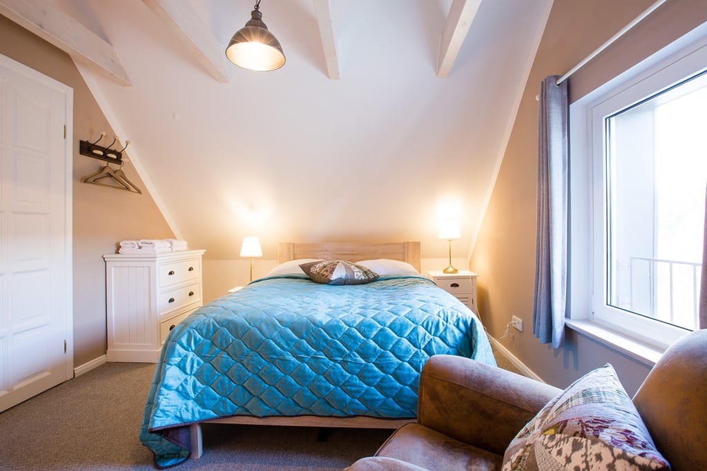 Schlafzimmer mit Doppelbett und Fenster, links eine Kommode, rechts ein Nachttisch mit Lampe, vorne ein Sessel.