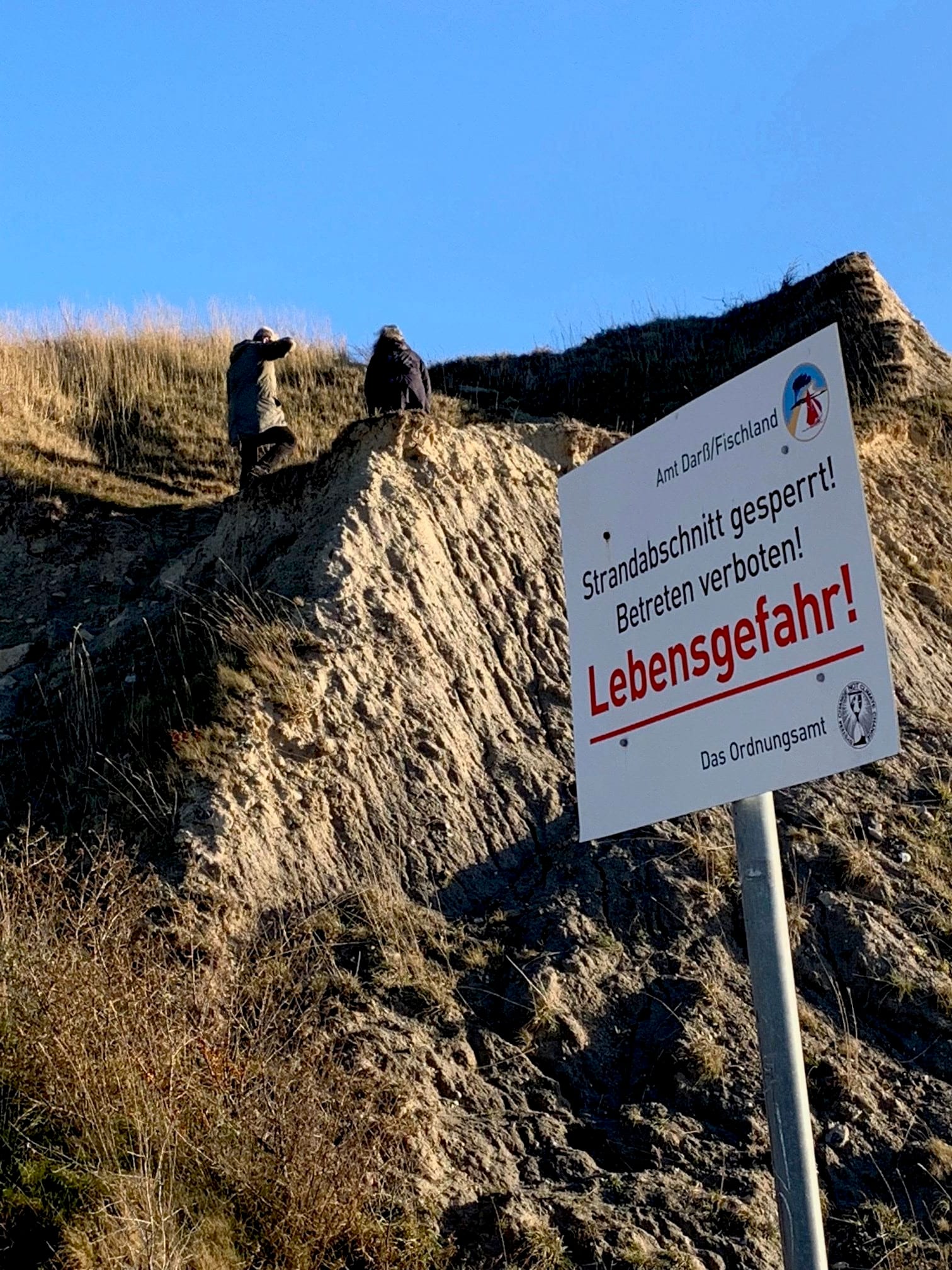 Schild: ‚Lebensgefahr – Betreten verboten’. Hinter dem Schild sind auf der Steilküste zwei Personen zu sehen.
