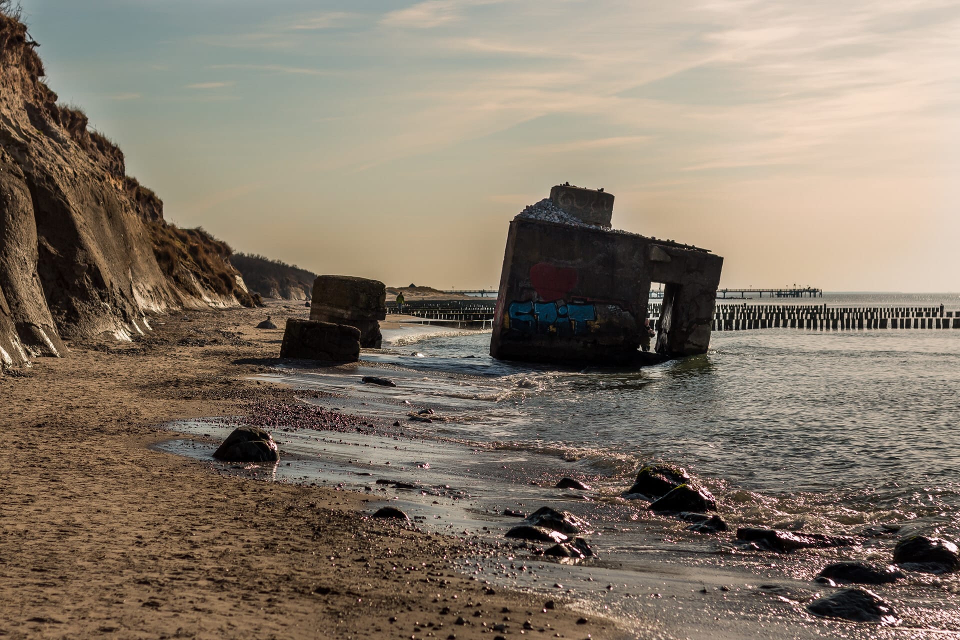 Blick vom Strandufer: links Steilküste, rechts im Wasser ein großes Betonelement und dahinter Buhnen.