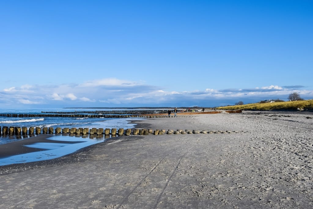 Ein weiter Sandstrand mit hölzernen Buhnen am blauen Meer unter sonnigem Himmel.