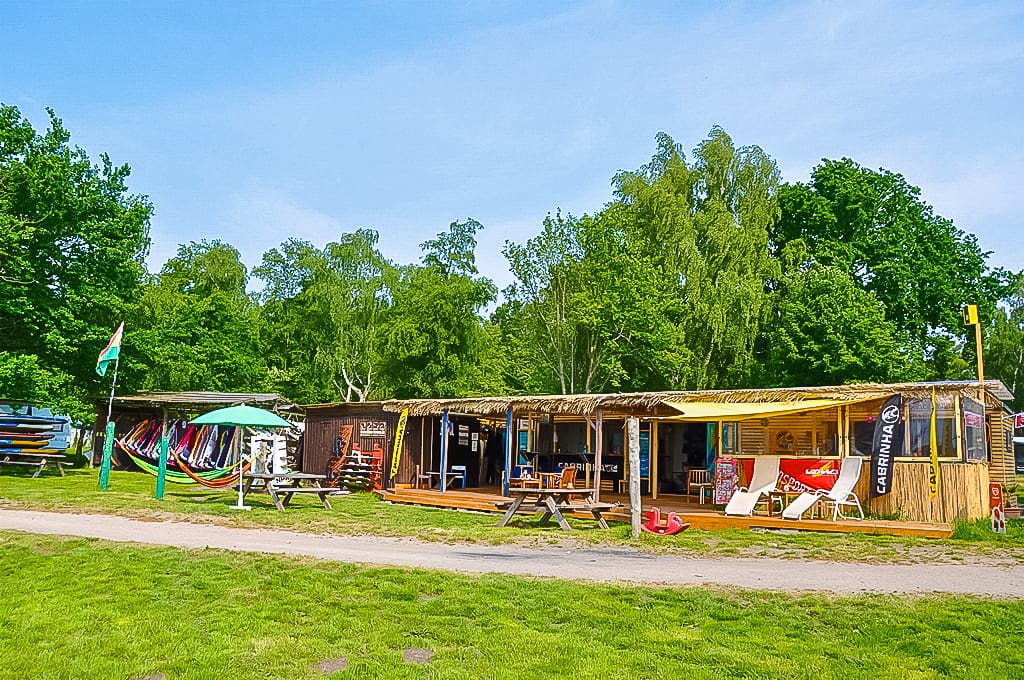 Ein hölzernes Gebäude mit Reetdach dient als Wassersportzentrum und lagert Ausrüstung wie Surfbretter und Segel in grüner Umgebung.