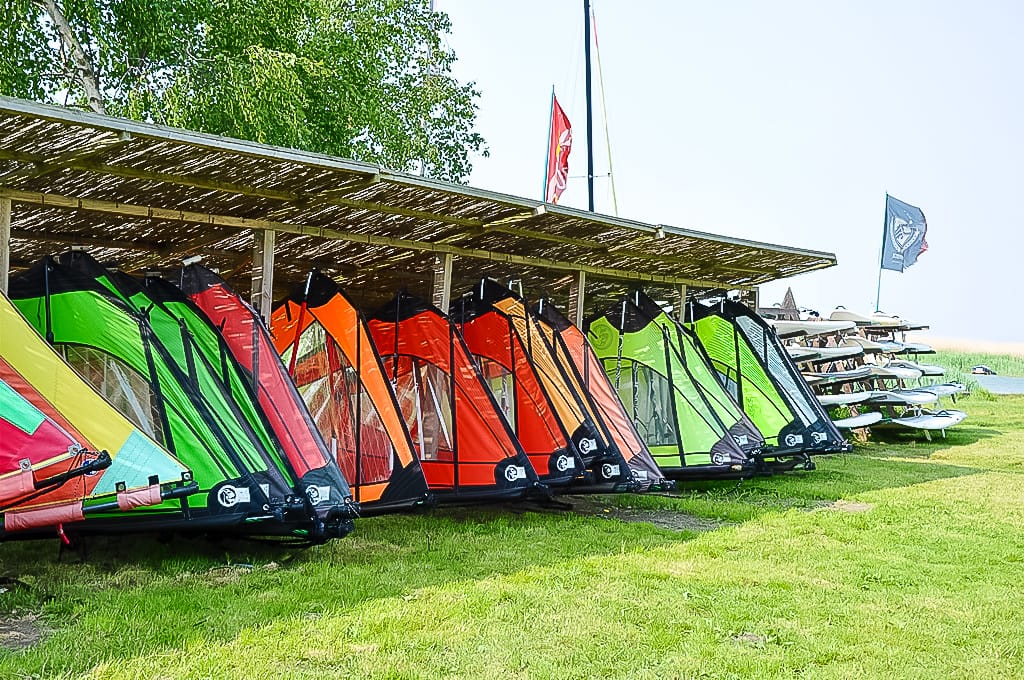 Mehrere bunte Windsurfbretter und Segel sind unter einem Reetdach in einem Wassersportzentrum gelagert.