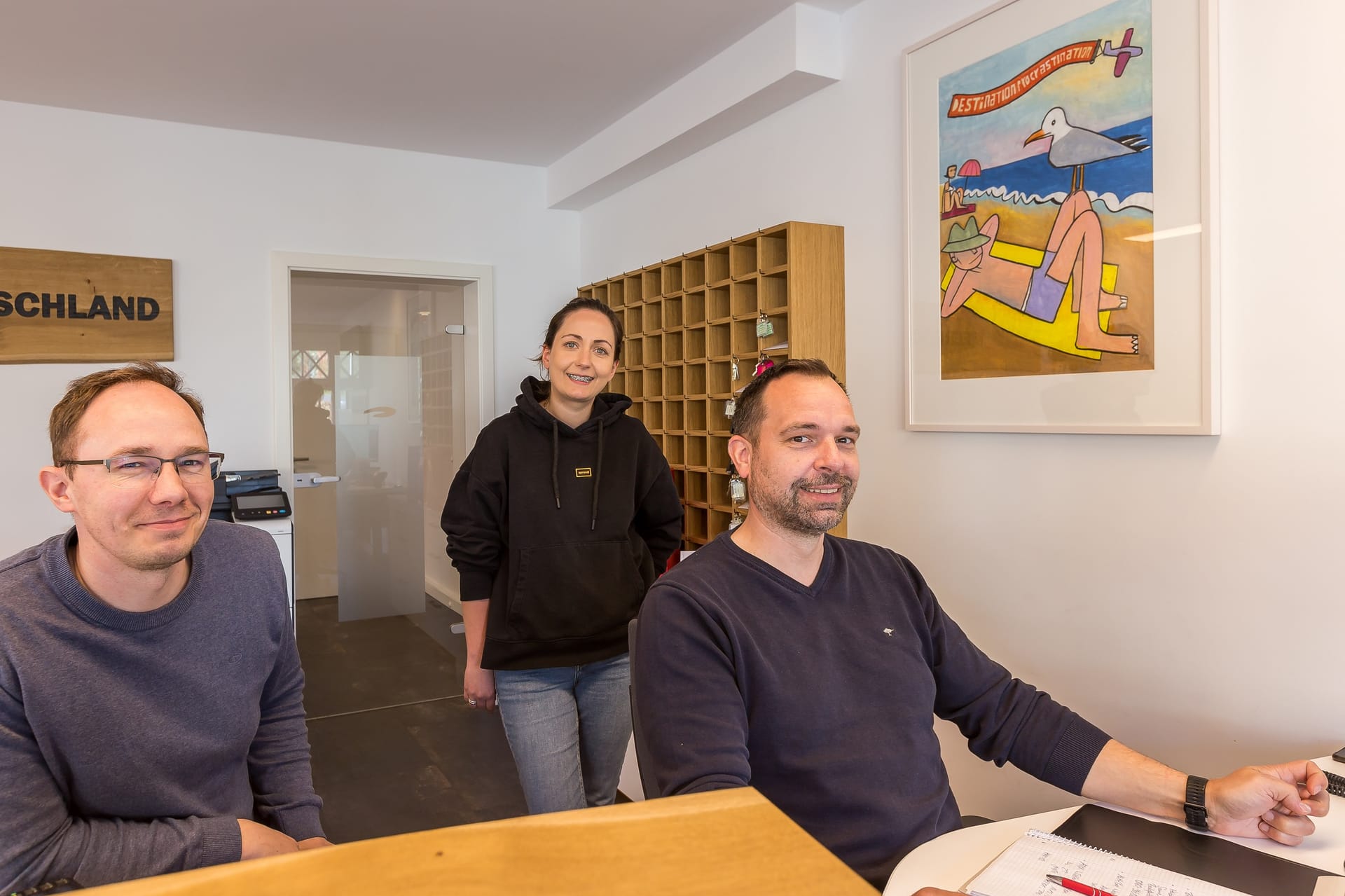 Agenturteam im Büro: Männer sitzen am Schreibtisch, Frau steht, alle lächeln.