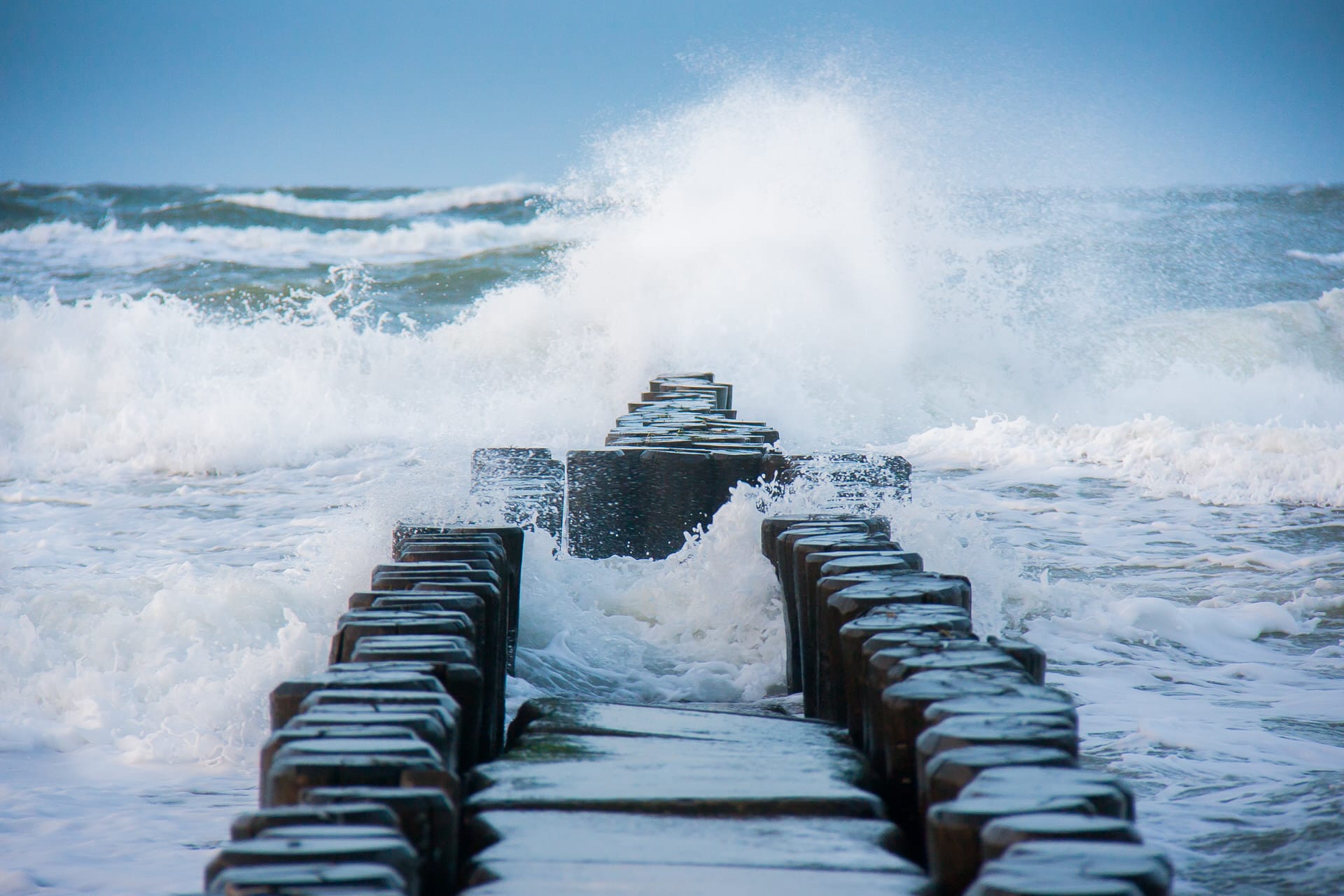 Holzsteg oder Buhne ins Meer hinein, Wellen brechen sich und spritzen Wasser hoch, bewölkter Himmel.
