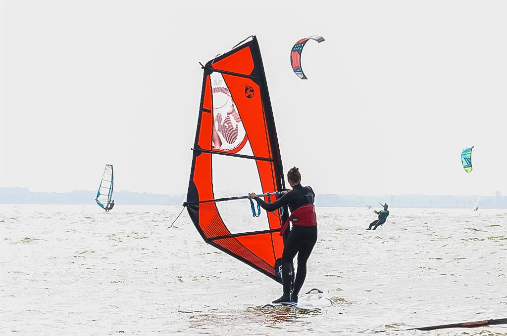 Eine Person mit Segel steht auf einem Surfbrett auf dem Wasser, umgeben von anderen Wassersportlern.