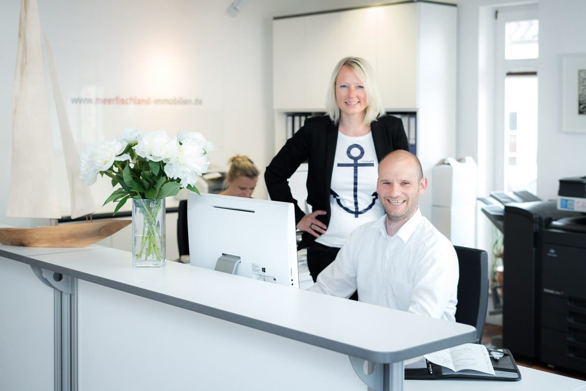 „Mitarbeiter im Büro: Eine Frau steht und lächelt, ein Mann sitzt am Computer und lächelt. Helle Einrichtung, im Hintergrund eine Frau am Schreibtisch.