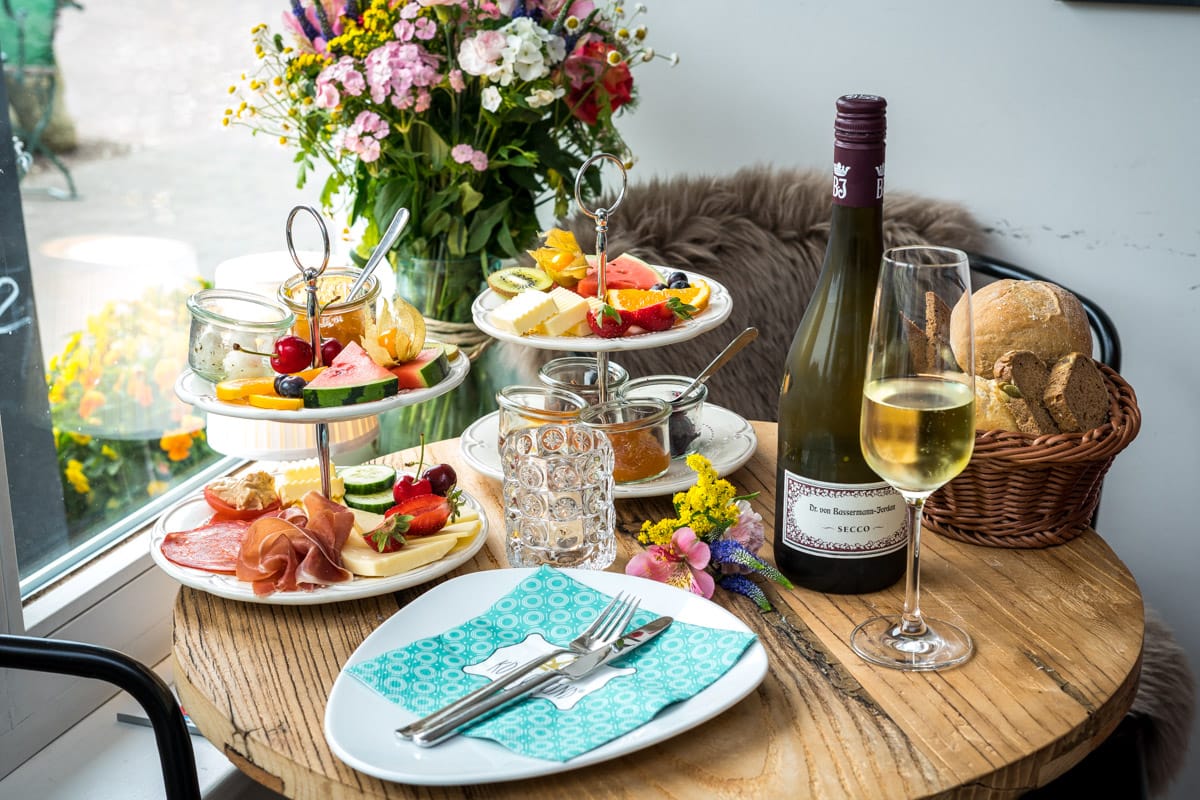 Ein gedeckter Tisch: Etagere mit Käse, Wurst und Früchten, Teller abgedeckt, Flasche Wein mit Glas, Brotkorb.