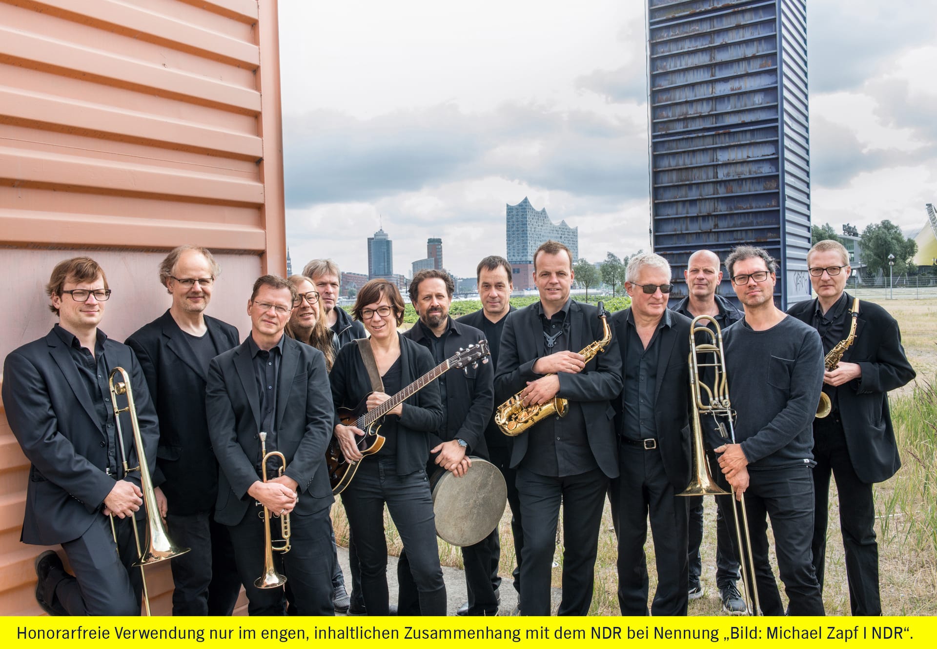 Eine Bigband posiert mit ihren Instrumenten vor einem braunen Gebäude und einem modernen Turm.