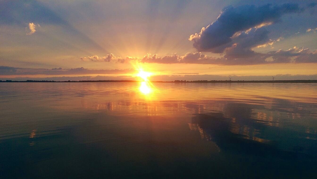 Die Sonne spiegelt sich bei Sonnenuntergang oder Sonnenaufgang auf einem Gewässer und sendet Lichtstrahlen durch die Wolken über dem Horizont.