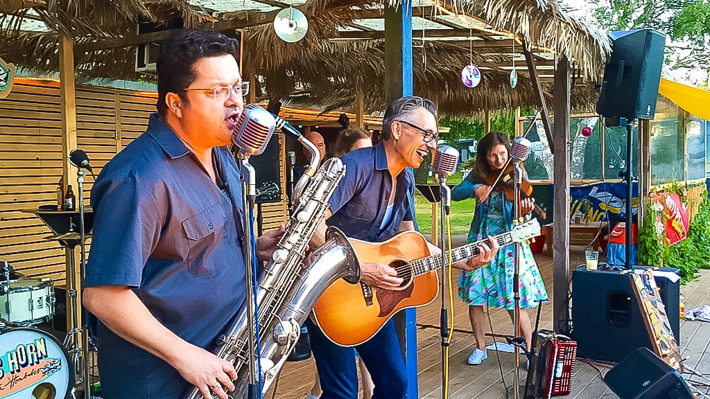 Zwei Männer spielen Saxophon und Gitarre in einer Band auf einer Bühne im Freien.