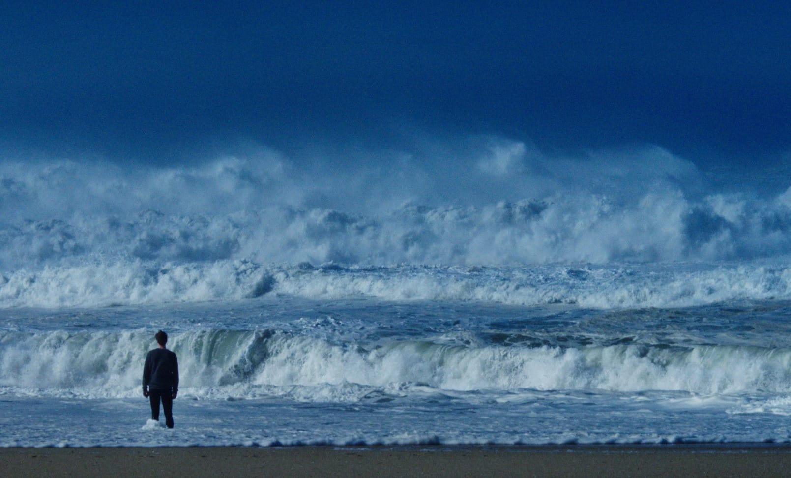 Ein Mann von hinten am Strand. Dunkle Kleidung. Er blickt auf das aufgewühlte Meer mit hohen, weißen Wellen. Dunkelblauer, wolkenloser Himmel.