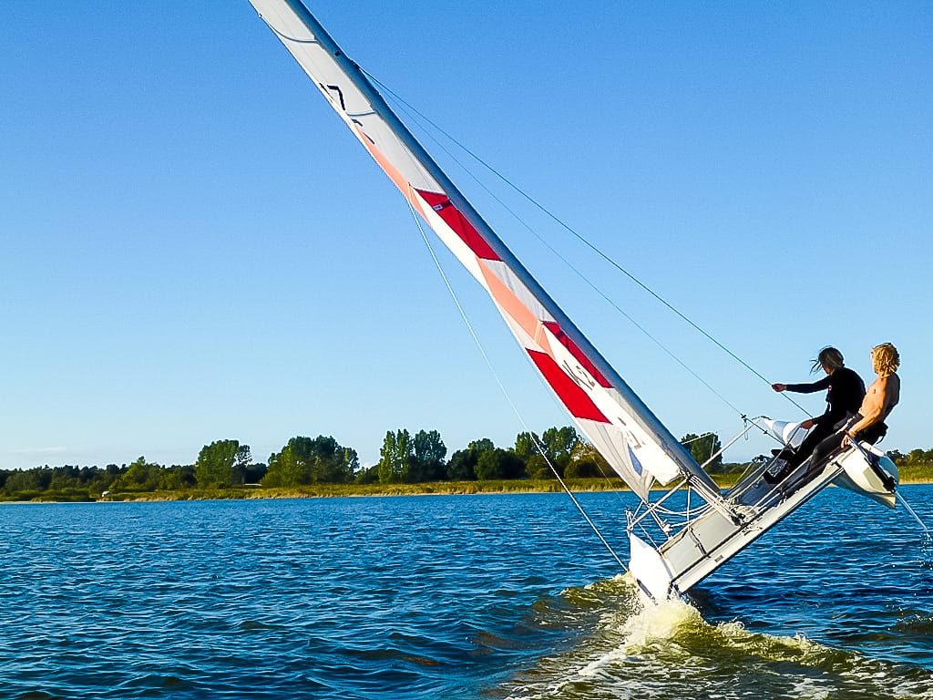 Zwei Personen segeln ein kleines Boot auf blauem Wasser mit einem Ufer im Hintergrund.