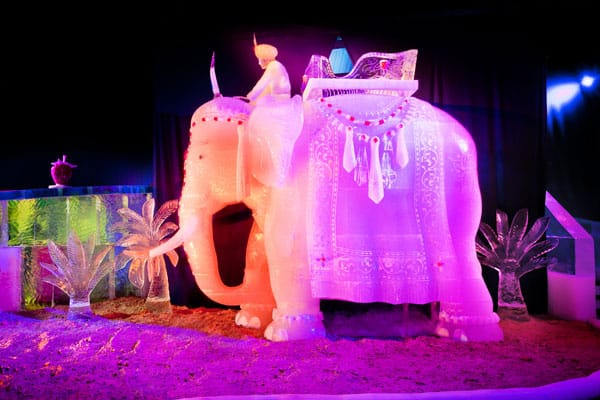 Eisskulptur eines indischen Elefanten mit Reiter. Die Figur ist mit einer verzierten Decke und Kopfschmuck versehen. Sie wird von innen in Rosa- und Orangetönen beleuchtet.