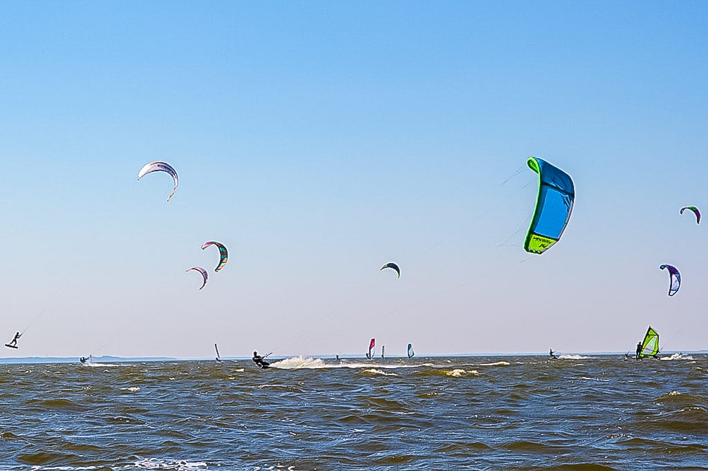 Mehrere Kitesurfer und Windsurfer sind auf einem Gewässer unter einem klaren, blauen Himmel aktiv.
