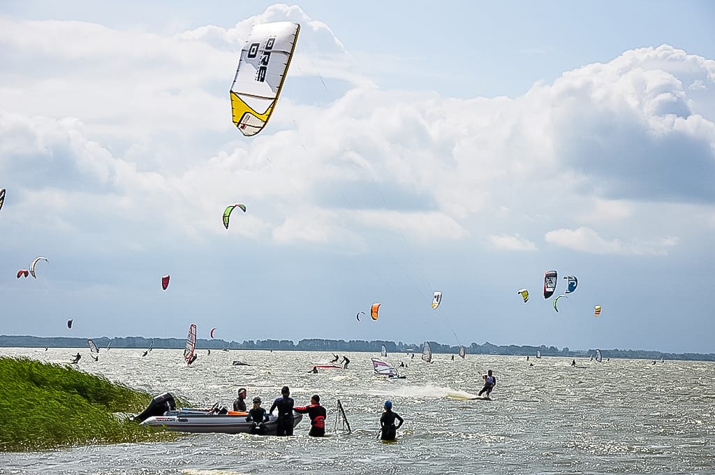 Auf einem Gewässer sind viele Menschen mit Kitesurfen und Windsurfen beschäftigt, während einige Personen in einem Boot am Rand des Schilfs stehen.