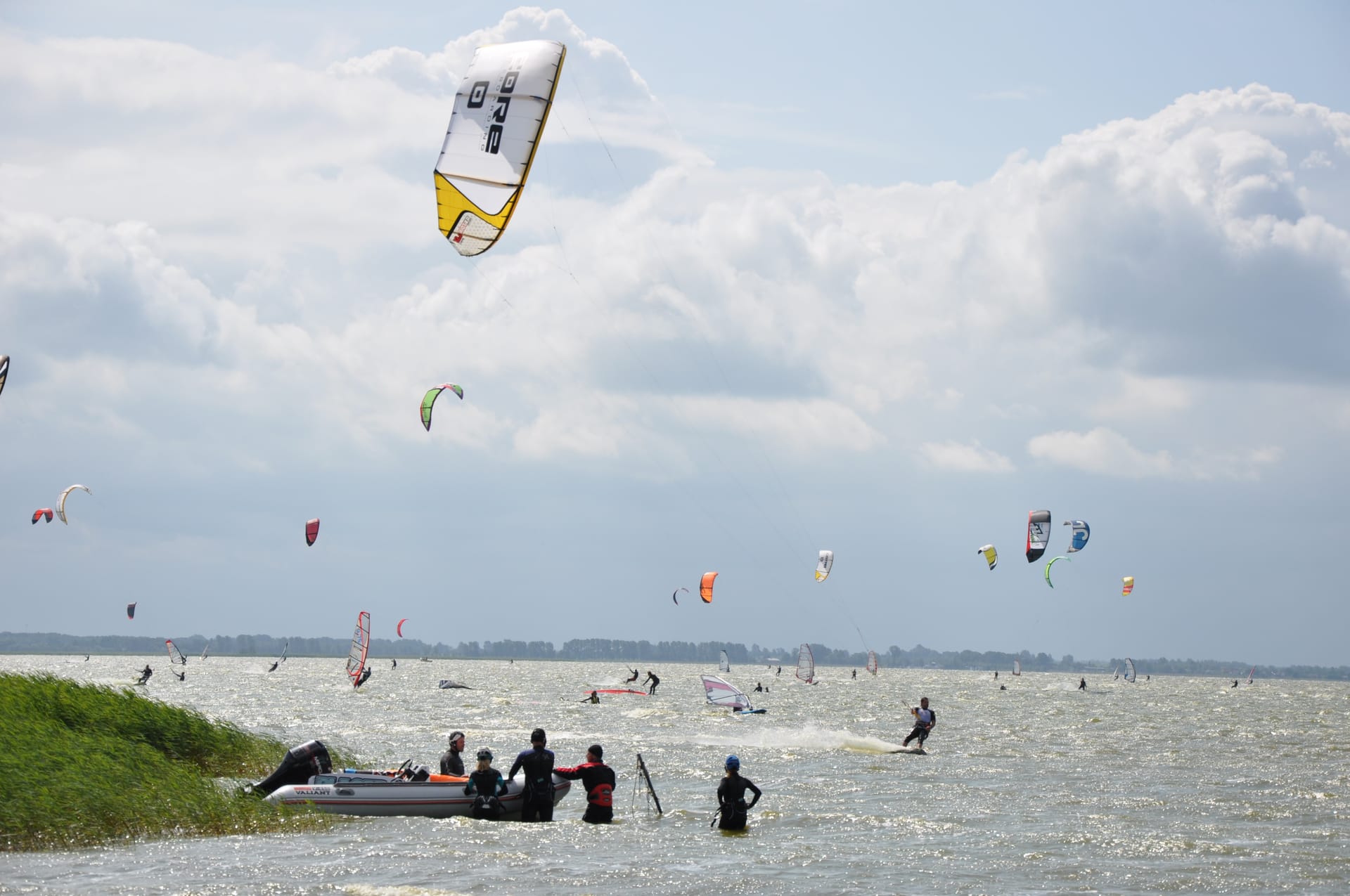 Auf einem Gewässer sind viele Menschen mit Kitesurfen und Windsurfen beschäftigt, während einige Personen in einem Boot am Rand des Schilfs stehen.