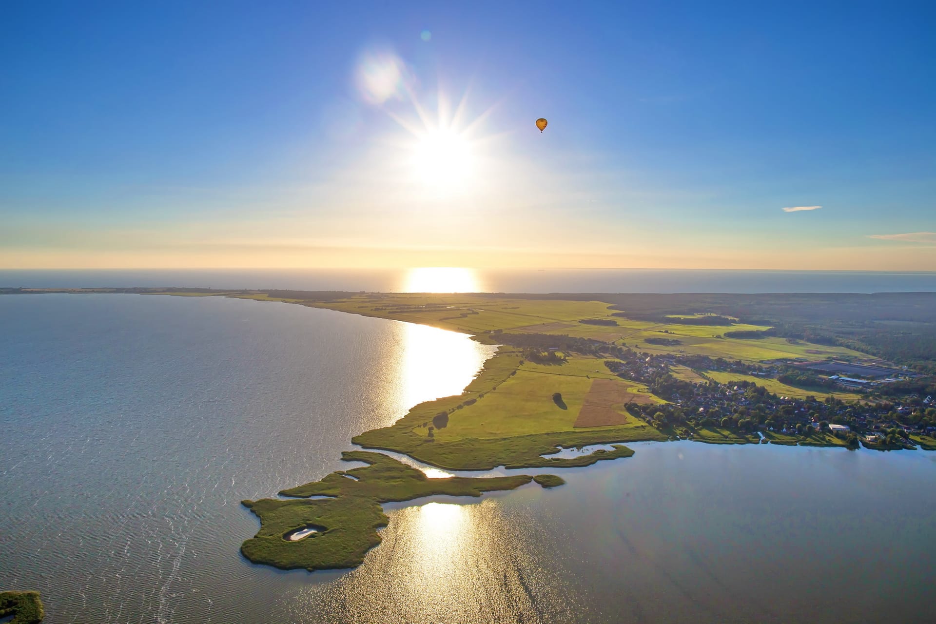 Luftaufnahme einer Boddenlandschaft an der Ostsee mit glitzerndem Wasser, grünen Wiesen, tief stehender Sonne und einem Heißluftballon am Himmel.