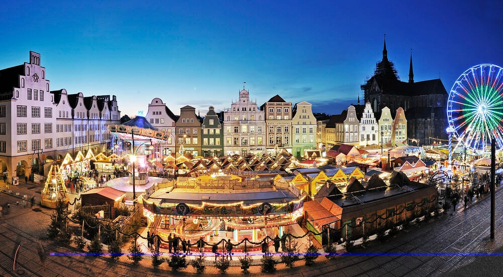 Ein Weihnachtsmarkt mit beleuchteten Ständen und Fahrgeschäften. Im Hintergrund stehen historische Gebäude und ein beleuchtetes Riesenrad.