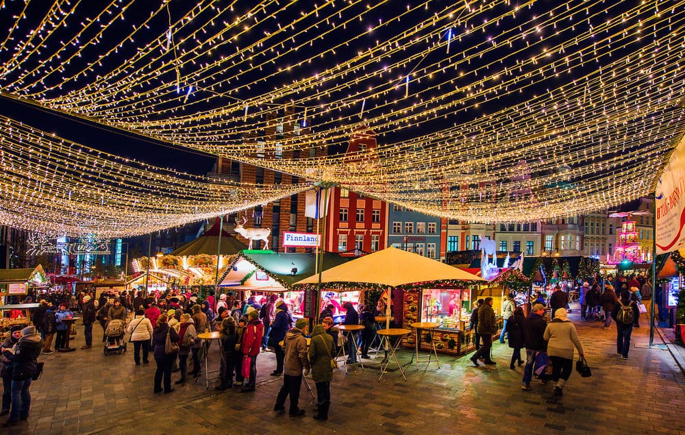 Auf dem Weihnachtsmarkt stehen die Menschen an den Ständen, unterhalten sich, essen und trinken, viele bummeln umher. Die Stände sind beleuchtet.