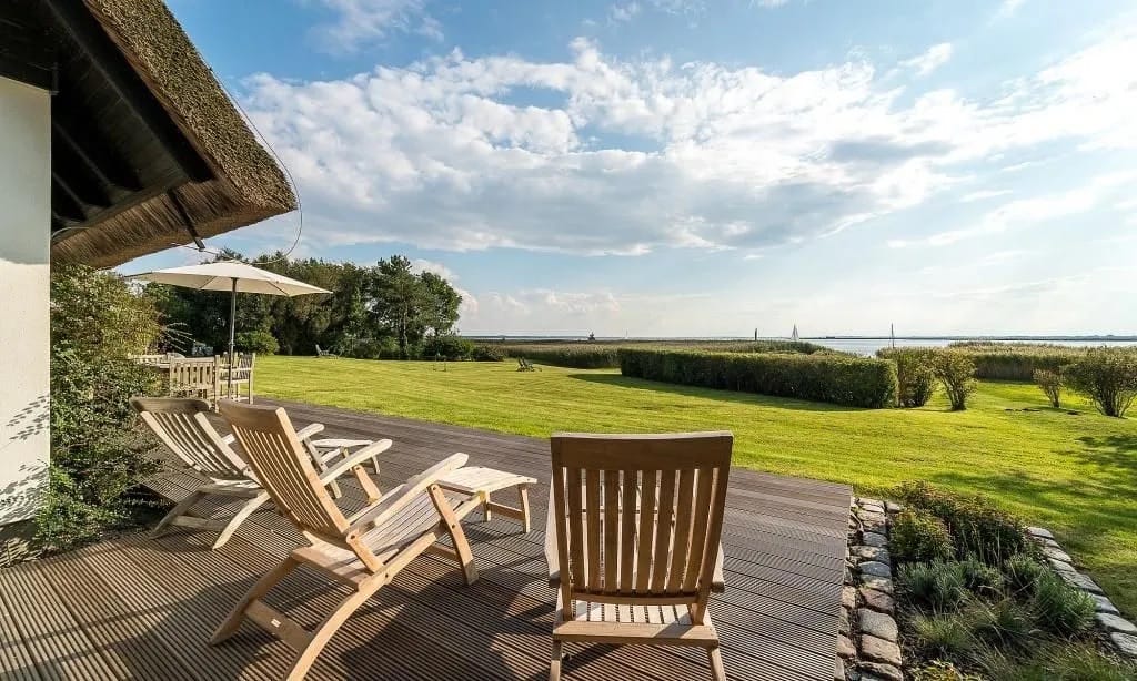 Holzterrasse mit Liegestühlen an Reetdachhaus mit Blick auf einen Rasen und das Meer.