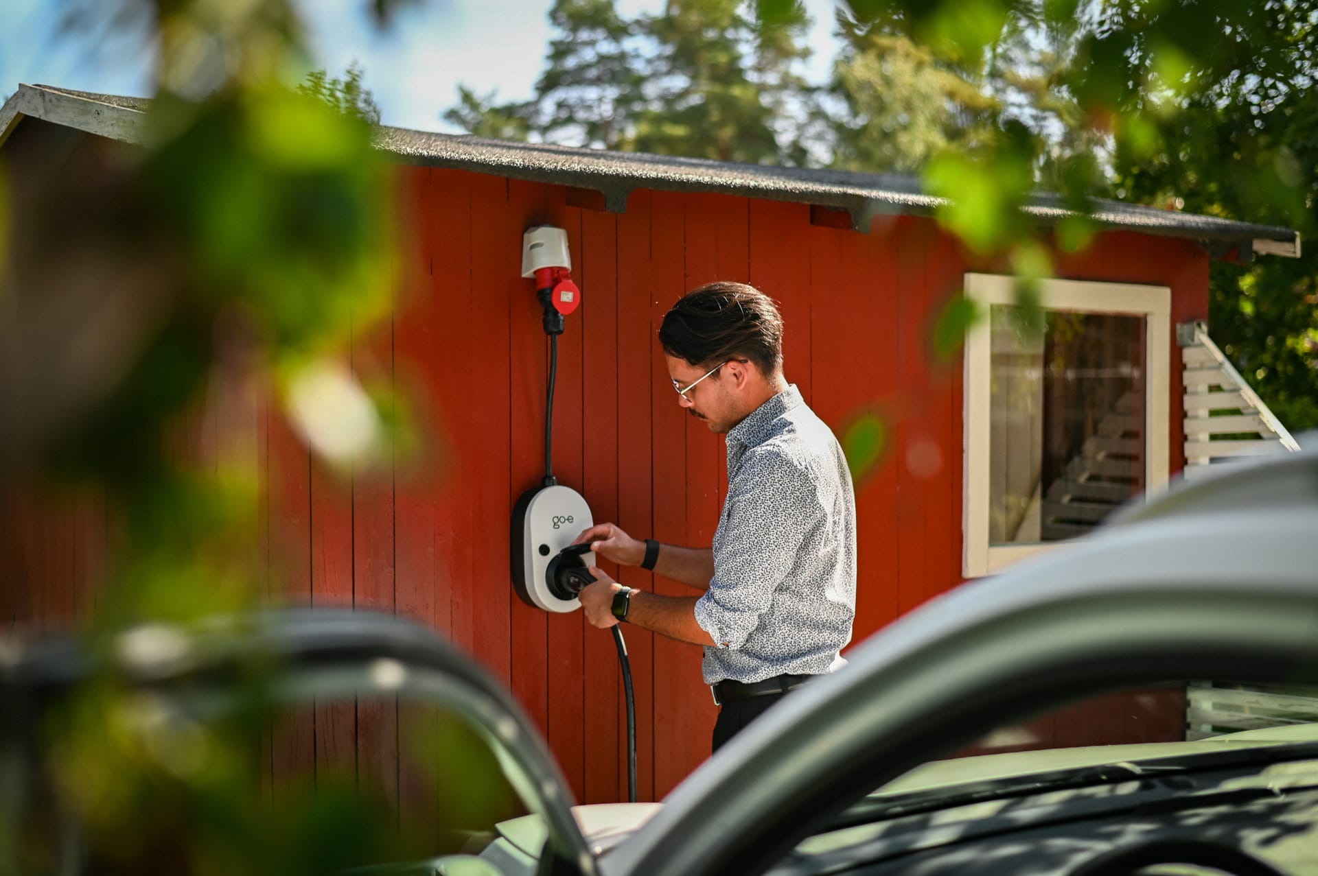 Mann schließt ein Ladekabel an eine go-e Wallbox an einem roten Holzhaus an.